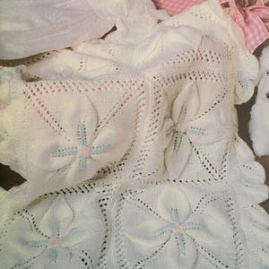 May include: A white crocheted baby blanket with a delicate floral pattern. The blanket features a repeating design of four-petal flowers with blue and pink accents.