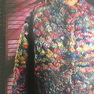 Op de afbeelding: Een veelkleurige gebreide cardigan met knoopsluiting. De cardigan is gemaakt met verschillende garens in tinten blauw, groen, paars, roze, geel en oranje.