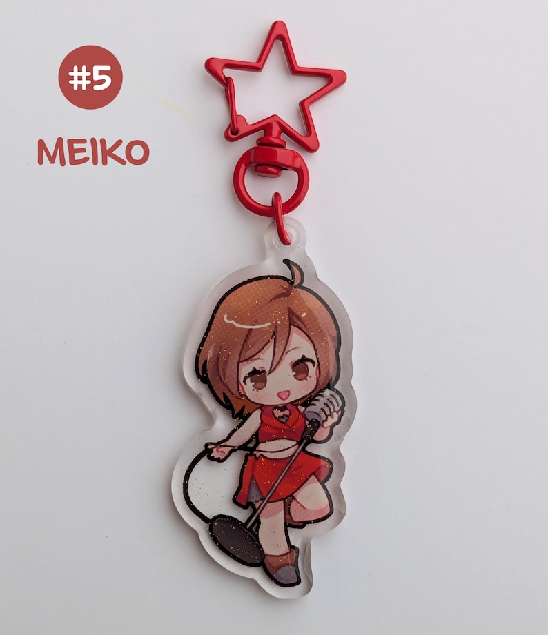 May include: A clear acrylic keychain featuring a cartoon character in a red outfit holding a microphone. The keychain has a red star-shaped clasp and the text "MEIKO" and "#5" in the upper left corner.