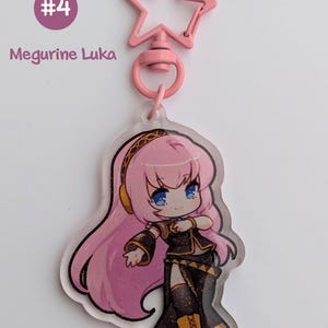 May include: A pink star-shaped keychain clip attached to a clear acrylic charm. The charm features a cartoon illustration of a character with pink hair, blue eyes, and a black and gold outfit. Text on the charm reads "Megurine Luka" and "#4".