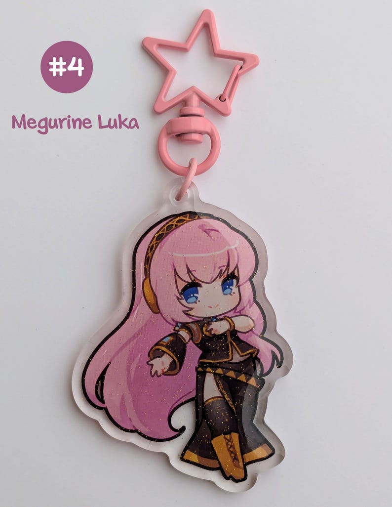 May include: A pink star-shaped keychain clip attached to a clear acrylic charm. The charm features a cartoon illustration of a character with pink hair, blue eyes, and a black and gold outfit. Text on the charm reads "Megurine Luka" and "#4".