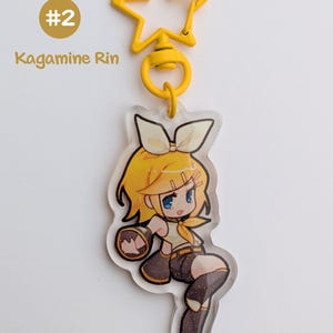 May include: A yellow star-shaped keychain with a cartoon character illustration. The character has yellow hair, a bow, and is wearing a yellow top and brown shorts. The text "#2 Kagamine Rin" is visible.