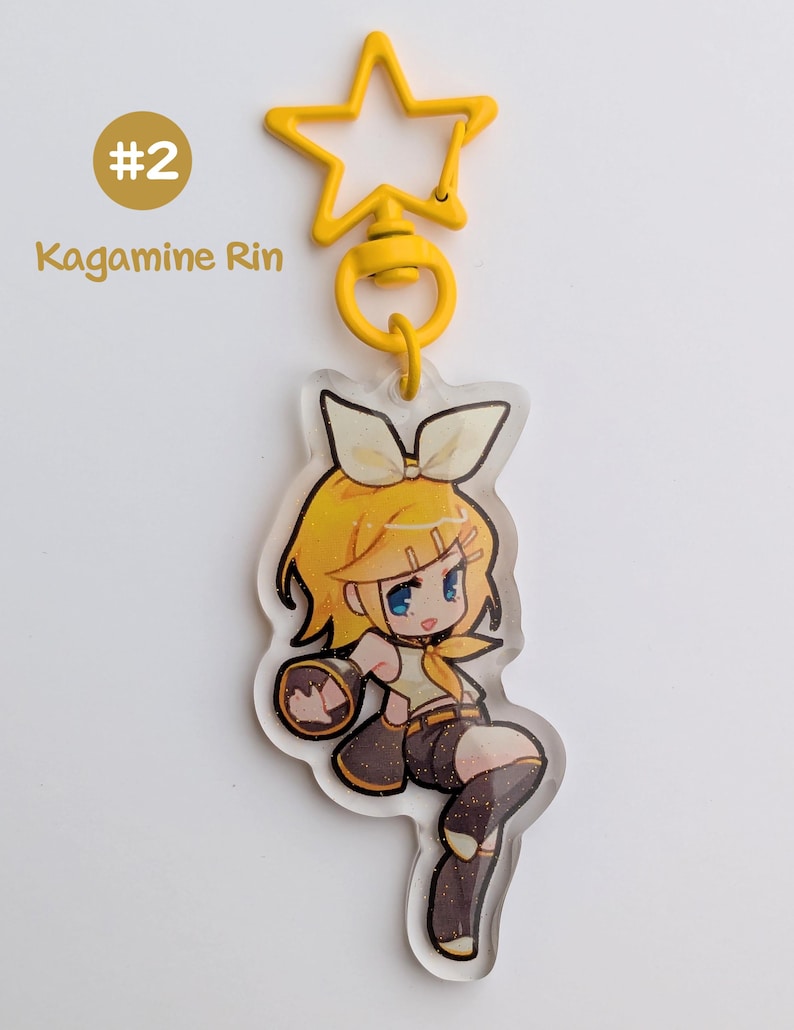 May include: A yellow star-shaped keychain with a cartoon character illustration. The character has yellow hair, a bow, and is wearing a yellow top and brown shorts. The text "#2 Kagamine Rin" is visible.