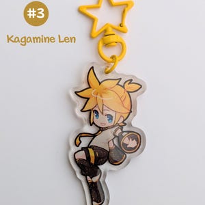 May include: A yellow star-shaped keychain with a cartoon character illustration. The character has yellow hair and blue eyes, wearing a black and gold outfit. The text "#3 Kagamine Len" is visible.