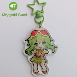 May include: A clear acrylic keychain featuring a cartoon character with green hair and red sunglasses. The character is wearing a green and orange outfit. The keychain has a green star-shaped clasp and the text "#7 Megpoid Gumi".