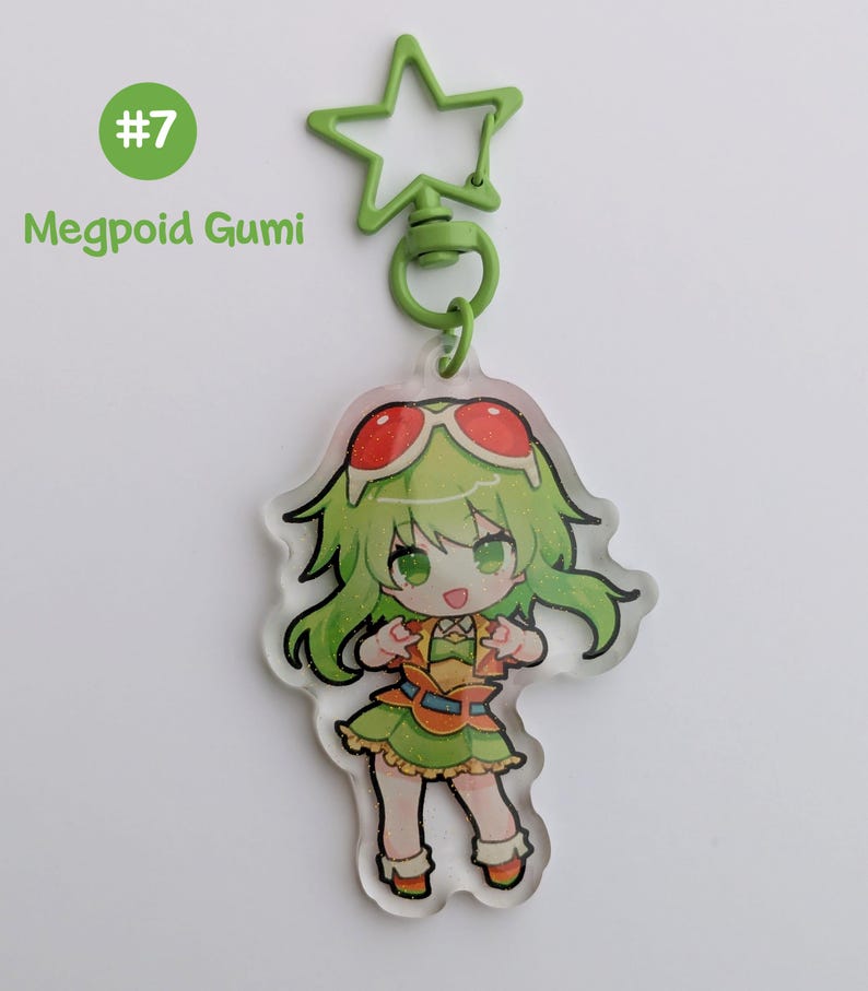 May include: A clear acrylic keychain featuring a cartoon character with green hair and red sunglasses. The character is wearing a green and orange outfit. The keychain has a green star-shaped clasp and the text "#7 Megpoid Gumi".