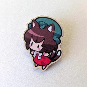May include: Wooden pin featuring a cartoon character with brown hair, cat ears, and a red dress. The character has a teal hat and a small gold hoop earring. The pin is cut in the shape of the character.