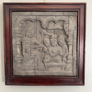 May include: A framed, gray stone relief carving depicting figures in a seated scene. The dark brown wooden frame surrounds the carved stone panel, which is suspended from a brass hook. The relief carving features detailed figures and architectural elements.