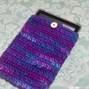 May include: A handmade crochet tablet sleeve in shades of purple, blue, and pink. The sleeve has a button closure and is designed to fit a tablet. The yarn has a textured appearance, and the overall design is simple and functional.