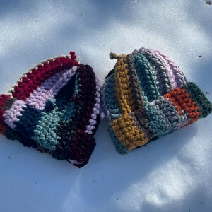 May include: Two handmade crochet beanies in a snowy setting. One beanie features a color block design with shades of red, purple, teal, and black. The other beanie has a similar design with mustard yellow, orange, green, and gray tones.