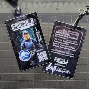 Può includere: Due tessere di identificazione in bianco e nero con una foto di un uomo e il testo "ACU" e "A DIVISION OF GEN SECURITY". Le tessere sono appese a un laccio nero con un fermaglio argentato.