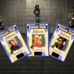 May include: Three plastic Jurassic Park iGen ID badges featuring Alan Grant, Ellie Sattler, and Ian Malcolm. Each badge has a blue and yellow design, the Jurassic Park logo, and a barcode.
