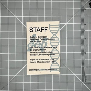 Op de afbeelding: Een witte plastic ID-kaart met een blauwe en grijze DNA-helixafbeelding. De kaart heeft de tekst "STAFF" in zwarte letters en bevat de tekst "Employee ID: 8675309", "Department: Parks and Recreation", "Site: Isla Sorna", "This card is Non-Transferable and is the property of INGEN. Its use is governed by the Ingen Employee Card Holder Agreement. Report lost or stolen cards to the Security Office at extension 4-001." en "INTERNATIONALGENETICTECHNOLOGIESINC".