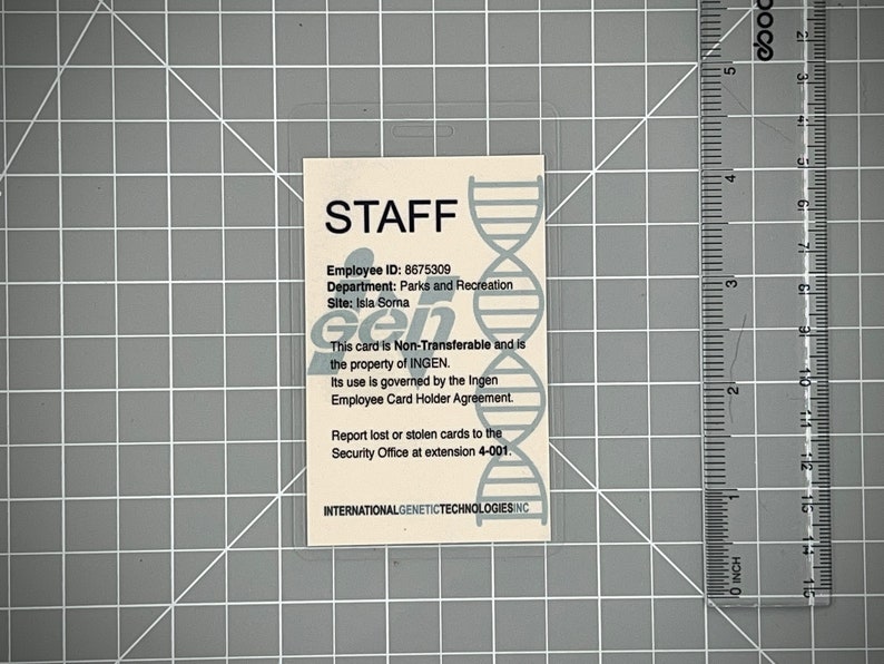 Op de afbeelding: Een witte plastic ID-kaart met een blauwe en grijze DNA-helixafbeelding. De kaart heeft de tekst "STAFF" in zwarte letters en bevat de tekst "Employee ID: 8675309", "Department: Parks and Recreation", "Site: Isla Sorna", "This card is Non-Transferable and is the property of INGEN. Its use is governed by the Ingen Employee Card Holder Agreement. Report lost or stolen cards to the Security Office at extension 4-001." en "INTERNATIONALGENETICTECHNOLOGIESINC".