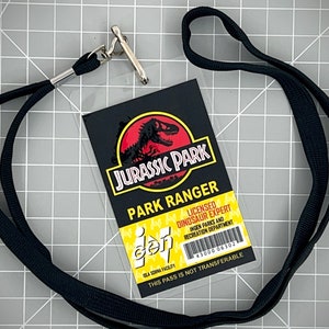 Op de afbeelding: Een zwarte lanyard met een zilveren clip houdt een doorzichtige plastic ID-kaart vast met een Jurassic Park-logo en de tekst "Park Ranger Licensed Dinosaur Expert" en "Ingen Parks and Recreation Department". De kaart heeft ook een barcode en de tekst "This pass is not transferable".