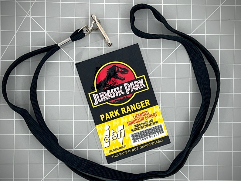 Op de afbeelding: Een zwarte lanyard met een zilveren clip houdt een doorzichtige plastic ID-kaart vast met een Jurassic Park-logo en de tekst "Park Ranger Licensed Dinosaur Expert" en "Ingen Parks and Recreation Department". De kaart heeft ook een barcode en de tekst "This pass is not transferable".