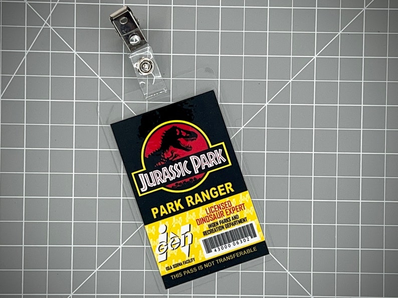Op de afbeelding: Een gele en zwarte Jurassic Park-ID-kaart met de tekst "Jurassic Park" en "Park Ranger". De kaart bevat ook het Jurassic Park-logo met een rood dinosaurus-silhouet. De tekst "Gecertificeerd dinosaurus-expert" en "Ingen Parks and Recreation Department" is ook inbegrepen. De kaart heeft een barcode en het nummer "43000 06302". De tekst "Deze pas is niet overdraagbaar" staat onderaan de kaart gedrukt.