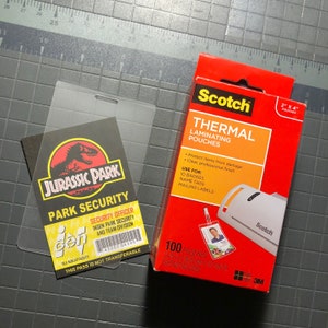 Op de afbeelding: Een transparante plastic hoes met een Jurassic Park beveiligingsbadge erin. De badge heeft een groen dinosaurus silhouet en de tekst "Jurassic Park Park Security". De hoes ligt naast een doos Scotch Thermal Laminating Pouches, die 5,1 cm bij 10,2 cm zijn en in een verpakking van 100 stuks zitten.