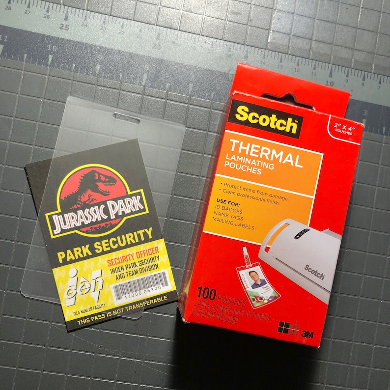 Op de afbeelding: Een transparante plastic hoes met een Jurassic Park beveiligingsbadge erin. De badge heeft een groen dinosaurus silhouet en de tekst "Jurassic Park Park Security". De hoes ligt naast een doos Scotch Thermal Laminating Pouches, die 5,1 cm bij 10,2 cm zijn en in een verpakking van 100 stuks zitten.