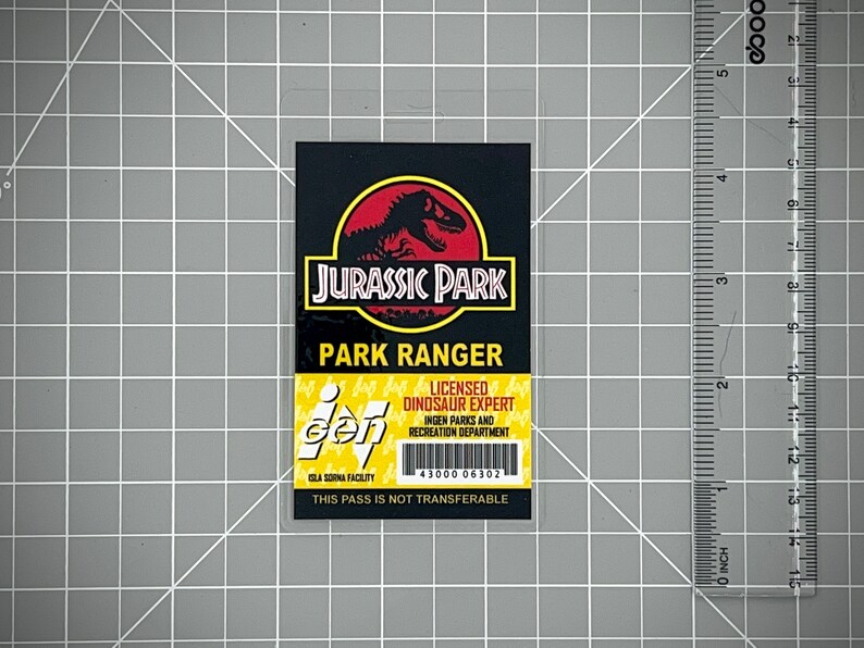 Op de afbeelding: Een gele en zwarte Jurassic Park park ranger ID-kaart met een zwart silhouet van een dinosaurus op een rode cirkel. De tekst op de kaart luidt "Jurassic Park Park Ranger Licensed Dinosaur Expert InGen Parks and Recreation Department Isla Sorna Facility 43000 06302 This Pass Is Not Transferable".