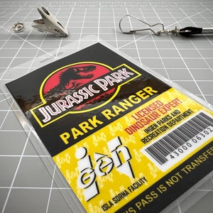 Op de afbeelding: Een gele en zwarte Jurassic Park-ID-kaart met de tekst "Jurassic Park Park Ranger" en "Gecertificeerd dinosaurusdeskundige". De kaart bevat ook de tekst "Isla Sorna Facility" en een barcode met het nummer "43000 06302". De tekst "US Pass is not transferable" staat onderaan de kaart gedrukt.