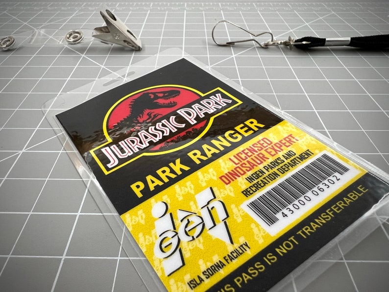 Op de afbeelding: Een gele en zwarte Jurassic Park-ID-kaart met de tekst "Jurassic Park Park Ranger" en "Gecertificeerd dinosaurusdeskundige". De kaart bevat ook de tekst "Isla Sorna Facility" en een barcode met het nummer "43000 06302". De tekst "US Pass is not transferable" staat onderaan de kaart gedrukt.