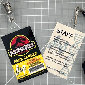Op de afbeelding: Twee plastic ID-kaarten met lanyards. De eerste kaart is een Jurassic Park Park Ranger-kaart met een zwarte achtergrond, een rood dinosauruslogo en de tekst "Jurassic Park Park Ranger". De tweede kaart is een witte personeelskaart met de tekst "STAFF" en "Employee ID: 8675309". Beide kaarten hebben de tekst "This pass is not transferable" en het logo "gen".
