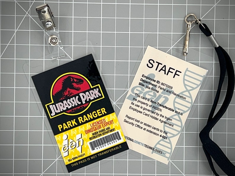 Op de afbeelding: Twee plastic ID-kaarten met lanyards. De eerste kaart is een Jurassic Park Park Ranger-kaart met een zwarte achtergrond, een rood dinosauruslogo en de tekst "Jurassic Park Park Ranger". De tweede kaart is een witte personeelskaart met de tekst "STAFF" en "Employee ID: 8675309". Beide kaarten hebben de tekst "This pass is not transferable" en het logo "gen".