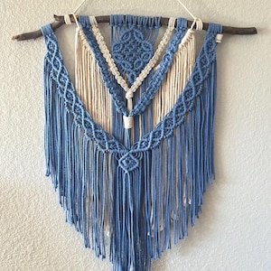 May include: A handcrafted macrame wall hanging featuring a natural wooden branch and intricate knotting. The design incorporates shades of blue and cream, with a geometric pattern at the top and cascading fringe. This decorative piece adds a bohemian touch to any space.