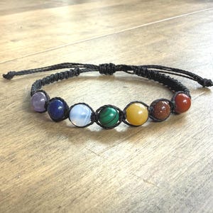 May include: A black macrame bracelet with seven gemstone beads in various colours. The beads are arranged in a row and include purple, blue, white, green, yellow, orange, and red stones. The bracelet is on a light brown wooden surface.