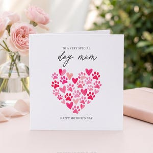 May include: A white greeting card with the text "TO A VERY SPECIAL dog mom" and "HAPPY MOTHER'S DAY". The card features a heart made of pink paw prints and hearts. Pink roses in a glass vase are in the background.