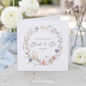 Puede incluir: Una tarjeta de felicitaci&oacute;n cuadrada blanca con una delicada corona de flores en acuarela en tonos rosa, azul y verde. El texto dice FOR THE BEAUTIFUL Bride to Be KELSEY con un peque&ntilde;o coraz&oacute;n rosa debajo, centrada sobre una superficie neutra.