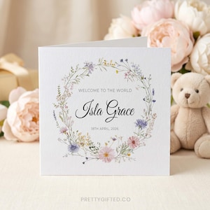 May include: A square white greeting card featuring a watercolor floral wreath in shades of pink, blue, and green. The text reads "WELCOME TO THE WORLD", "Isla Grace", and "18TH APRIL, 2026" in black ink. The card has a textured paper finish.
