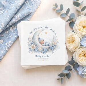 May include: A stack of square paper napkins featuring a sleeping baby in a blue hat resting on a crescent moon. The design includes blue and white floral accents, a gold star, and the text: We are over the moon, Baby Carter is on the way.