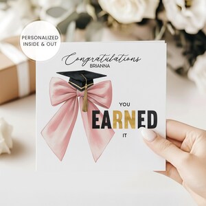May include: A graduation card with a pink bow and a graduation cap. The card says "Congratulations Brianna, You Earned It". The card is held by a person.