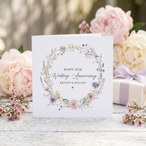 May include: A square white greeting card featuring a delicate floral wreath illustration in shades of pink, purple, and yellow. The text reads HAPPY 40TH WEDDING ANNIVERSARY KRISTEN & WILLIAM with a small red heart centered below the names.