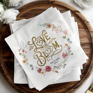 May include: White napkins with a floral design and gold lettering that reads "Love in Bloom" with the names "Kym & Mark" and the date "12.10.25".