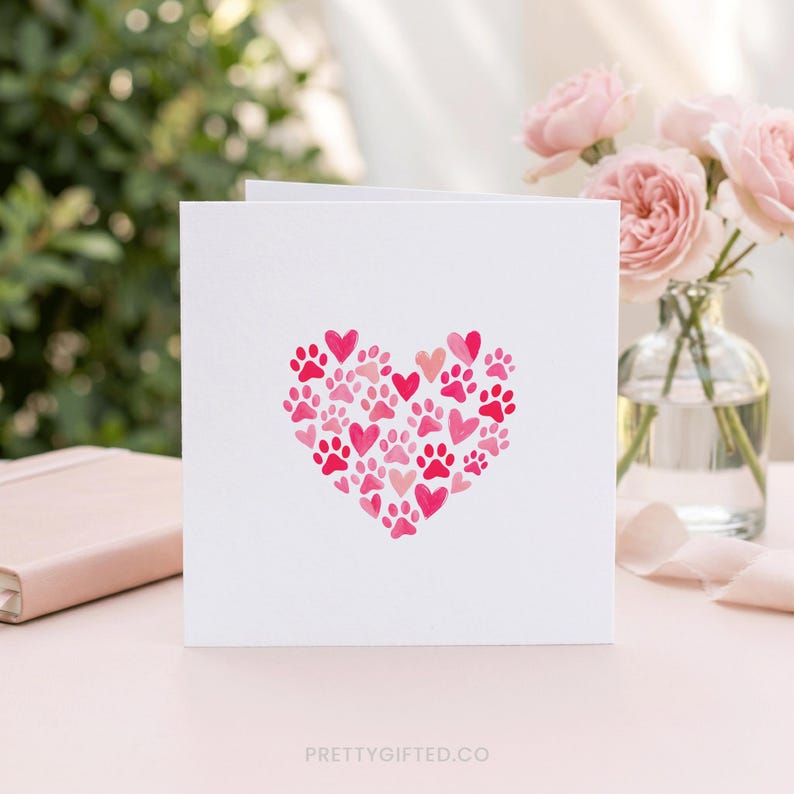 May include: White greeting card featuring a heart design made of pink paw prints and hearts. The card is displayed on a pink surface with a blurred background of greenery and flowers. The text "PRETTYGIFTED.CO" is at the bottom.