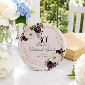 Peut inclure: Un ornement rond en céramique avec un fond rose pâle, des lignes géométriques dorées, des fleurs blanches et violet foncé, et le texte noir 30th Anniversary, Charlie & Sarah, July 28th, 1994 - 2024, suspendu par un cordon doré.