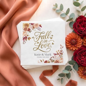 May include: A stack of square white paper napkins featuring gold cursive text that reads Fall in Love, with the names Katie & Mark and the date October 3rd, 2026 printed below. The design includes floral illustrations in shades of orange and brown.