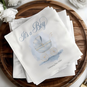 May include: A stack of white napkins with blue and gold accents. The top napkin features a watercolor illustration of a baby carriage and the text "It's a Boy" in blue cursive. The text "Celebrating Baby Miller" is printed in black below the illustration.