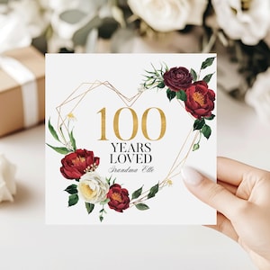 May include: A square card with a gold geometric heart design, featuring the text "100 YEARS LOVED Grandma Elle" in gold. The heart is adorned with burgundy and white flowers and green leaves. The card is held by a person.