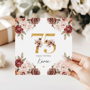 May include: A square birthday card with the number 75 in gold, adorned with floral arrangements. The card reads "Happy Birthday Karen" in elegant script, surrounded by roses and greenery. The card is held by a person.