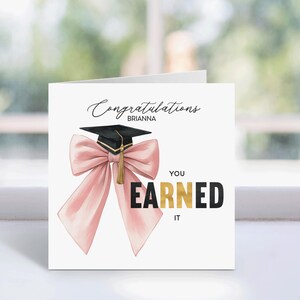 May include: A white graduation card with the text "Congratulations Brianna" in elegant script. A black graduation cap with a gold tassel is featured above a large pink bow. The words "YOU EARNED IT" are in black and gold.
