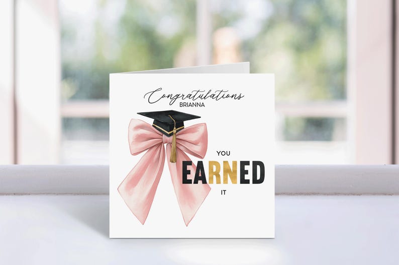 May include: A white graduation card with the text "Congratulations Brianna" in elegant script. A black graduation cap with a gold tassel is featured above a large pink bow. The words "YOU EARNED IT" are in black and gold.