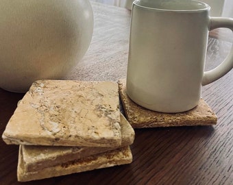 Op de afbeelding: Een stapel van vier vierkante, beige stenen onderzetters met een getextureerd oppervlak. Een witte mok staat op een onderzetter. Een vaas met gedroogde bladeren staat op een donkere houten tafel.
