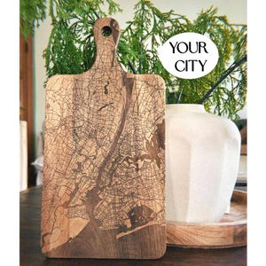 May include: Wooden chopping board with a city map design. The board has a handle with a hole and features the text "YOUR CITY" in a white circle. The board is displayed on a dark wooden table.