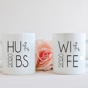 Puede incluir: Dos tazas de cerámica blanca con texto negro. La taza de la izquierda dice "2020 Hubs" y la taza de la derecha dice "2020 Wife".