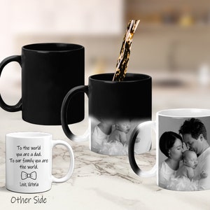 May include: Two black color-changing mugs with a white mug next to them. The white mug has a message that reads "To the world you are a dad. To our family you are the world. Love, Victoria". The black mugs have a photo of a family with a baby. One mug is filled with a brown liquid.