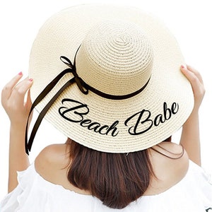 May include: A beige straw sun hat with a black ribbon and the text "Beach Babe" embroidered on the brim.